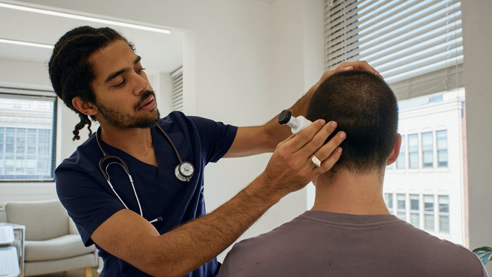 Doctor in navy scrubs examining the back of a man's head with a handheld magnifier in a bright consultation room