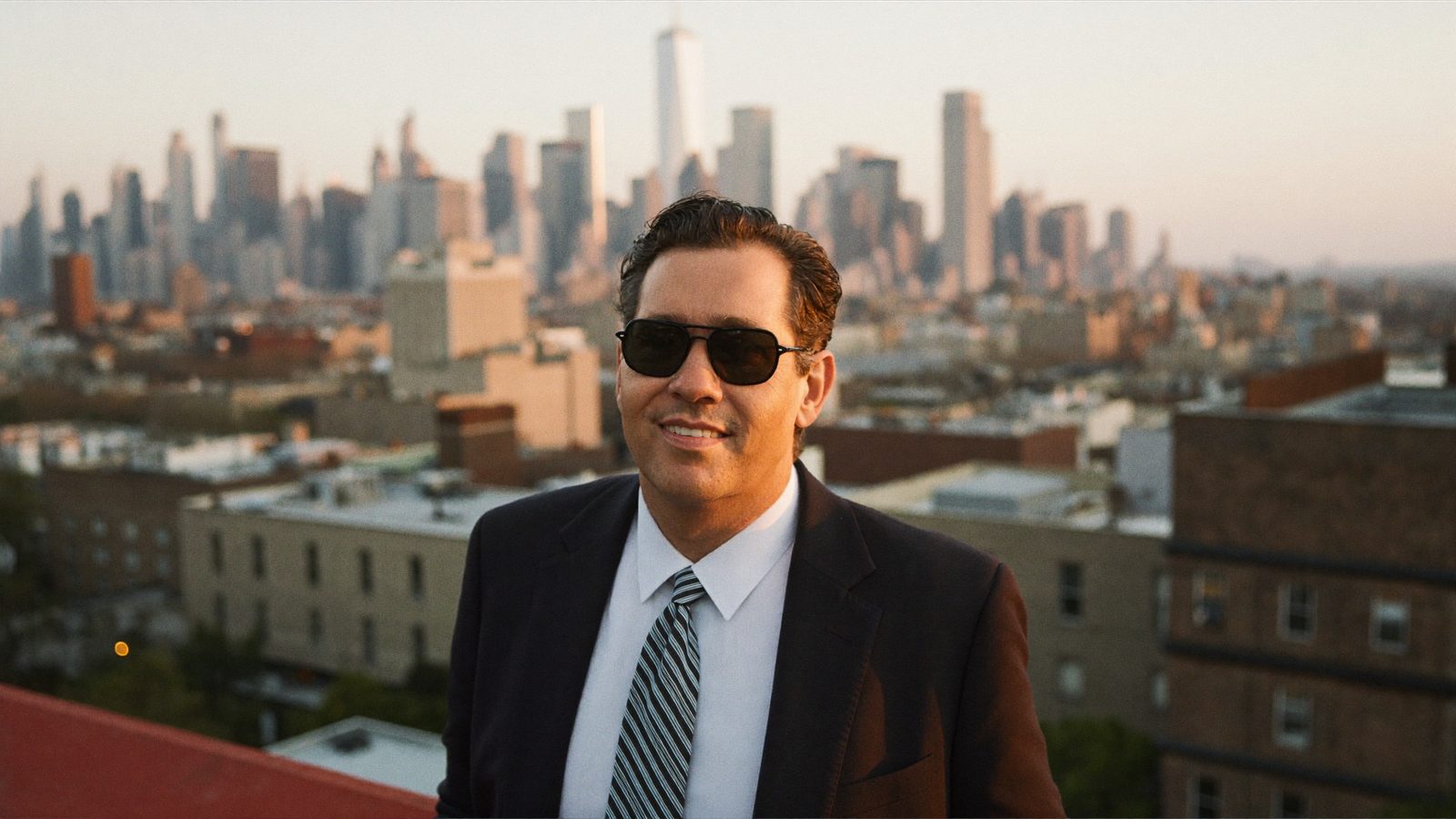 Smiling man in a suit and sunglasses on a Brooklyn rooftop at golden hour with the Manhattan skyline behind