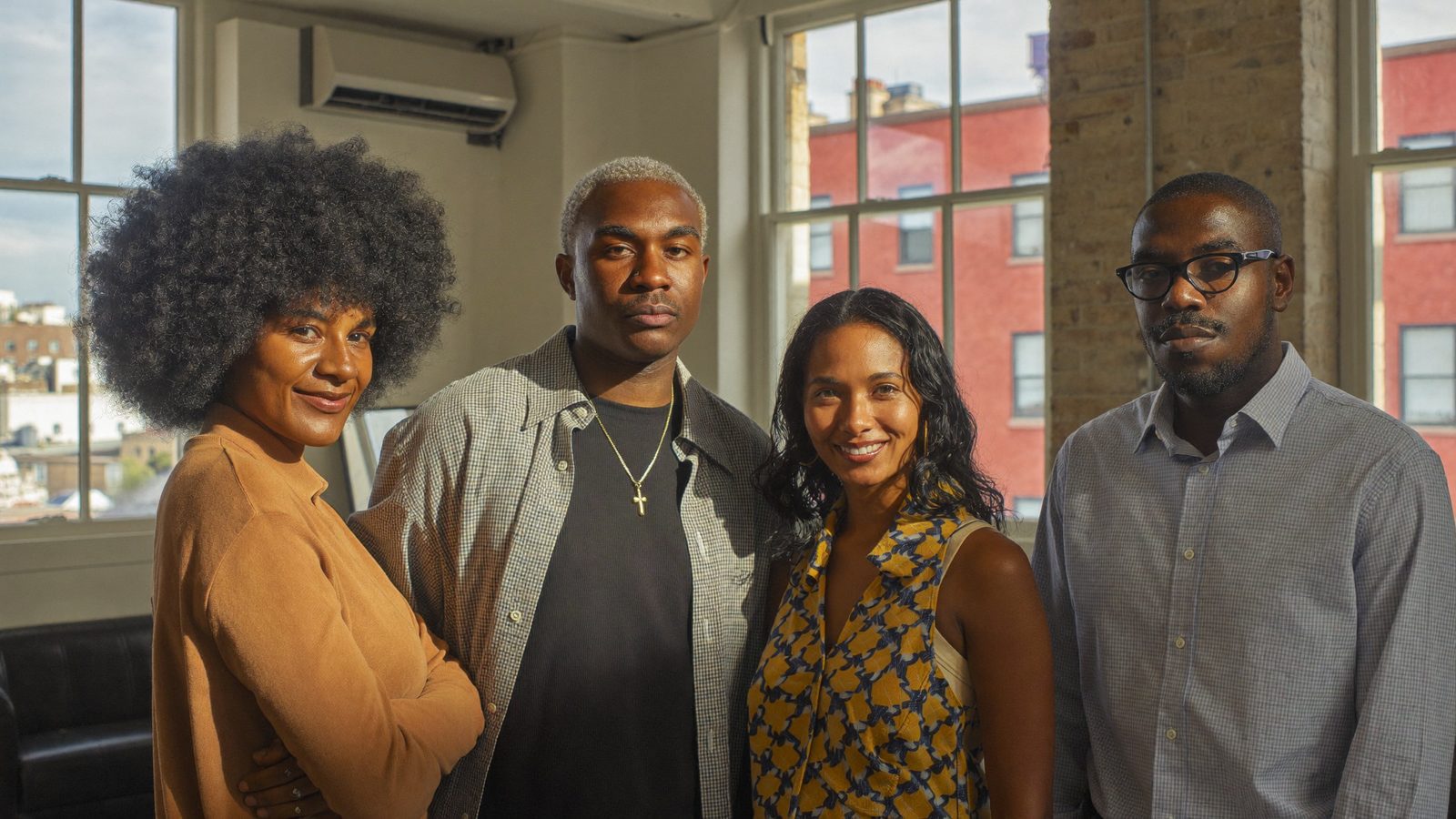 Four adults with different hair types, including a tall Afro and short dark hair, standing together in a sunlit loft