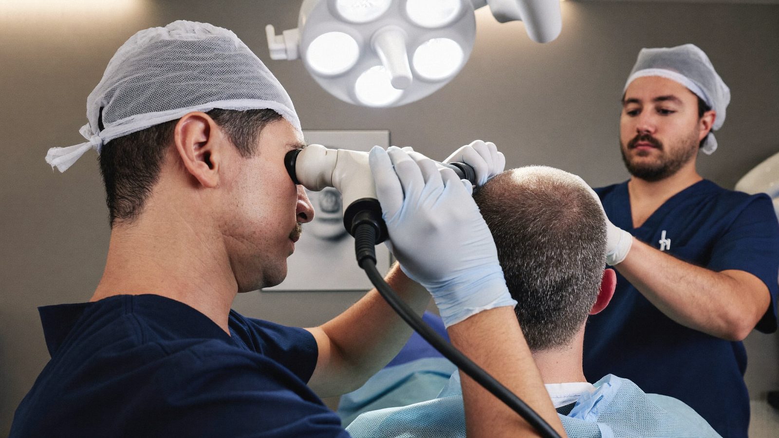 Surgical team member wearing magnification loupes working on the back of a patient's scalp while a doctor supervises