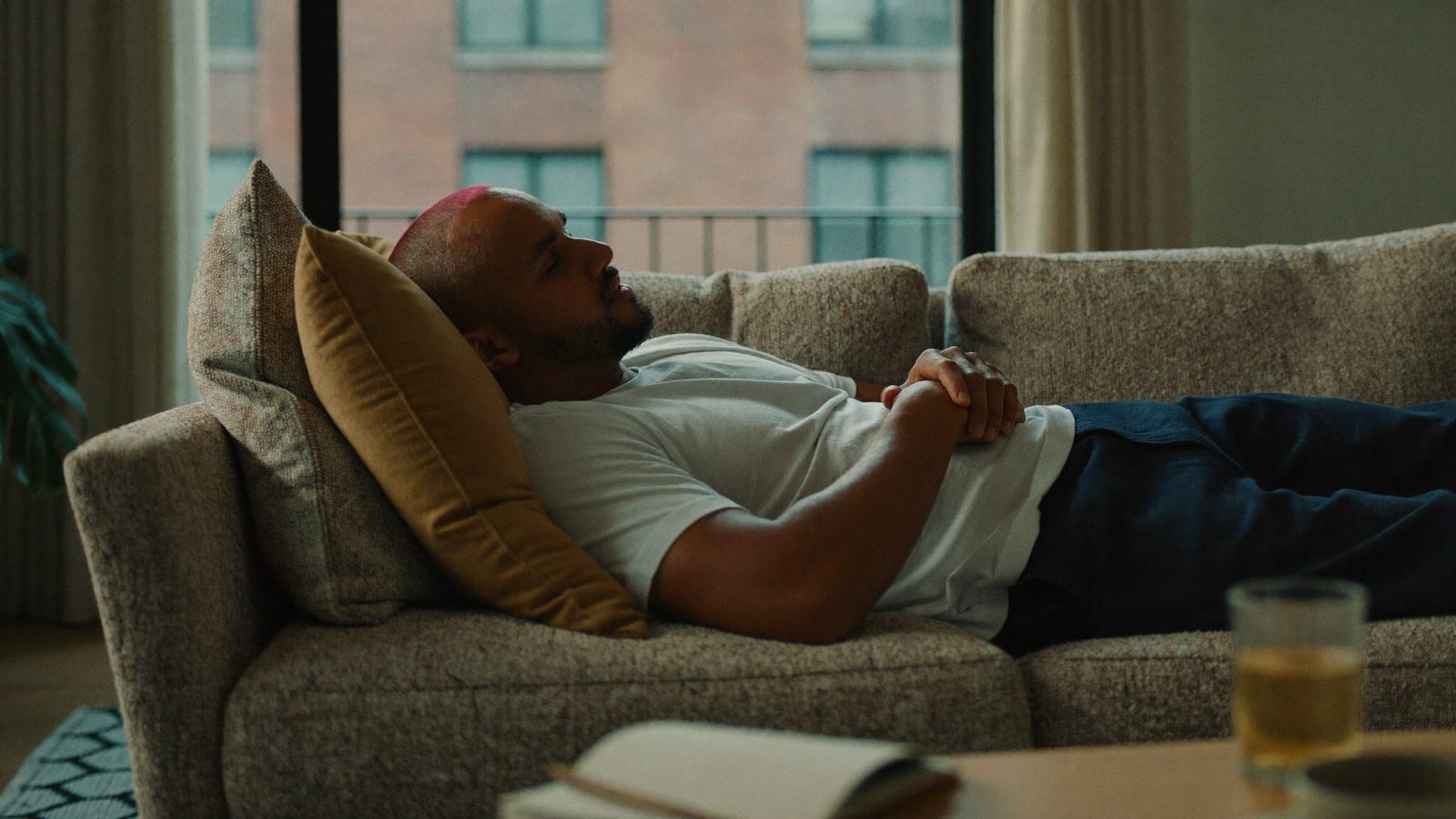 Man resting on a sofa at home with his head propped on pillows and a healing hairline, a drink and notebook on the table