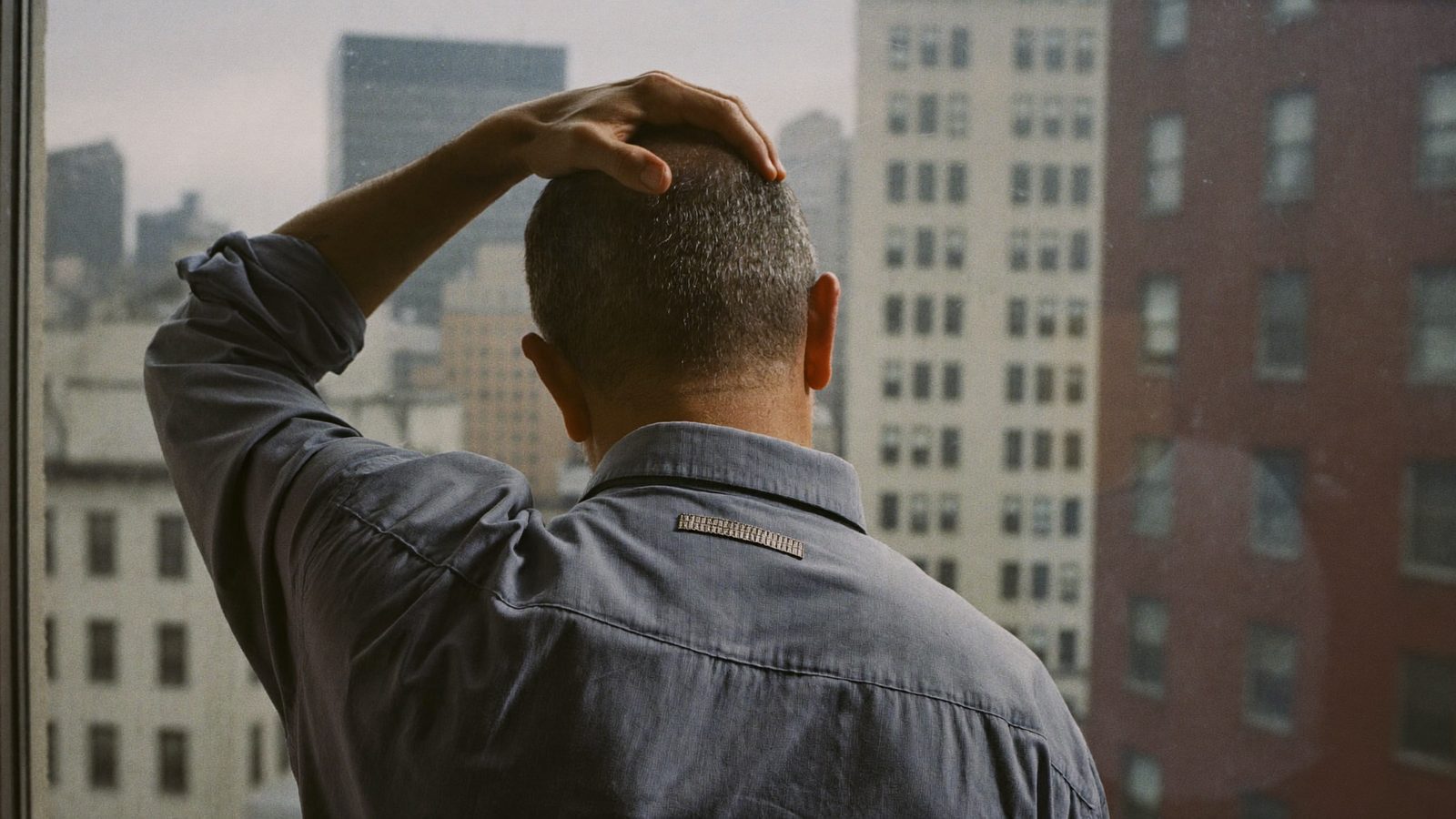 Man seen from behind running a hand over his thinning hair while looking out a window at city buildings